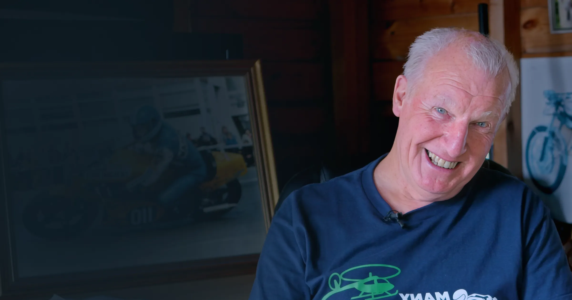 Smiling man in a dark blue shirt sits indoors with a motorbike photo in the background. The scene conveys warmth and nostalgia.